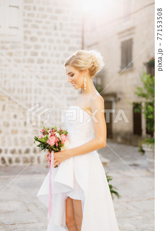 A bride stands on the street of the old town of Perast, holds a bouquet in her hands and smiles  A bride stands on the street of the old town of Perast, holds a bouquet in her hands and smiles  77153508
