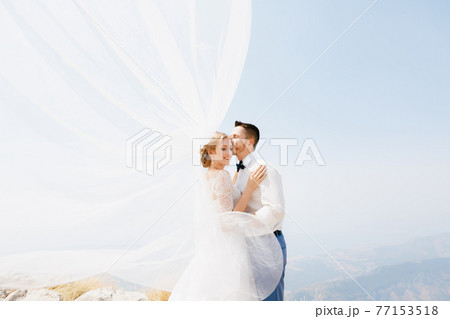 The bride and groom are embracing on the mountain, the groom kisses the bride, the wind waves the veil The bride and groom are embracing on the mountain, the groom kisses the bride, the wind waves the veil 77153518