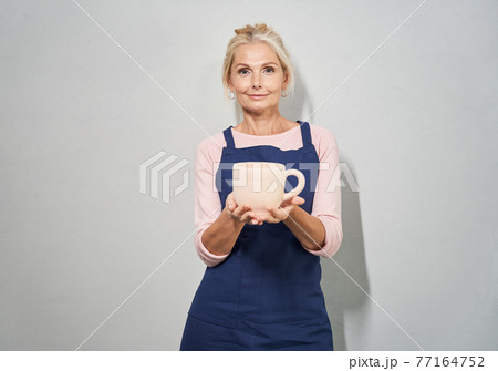 Portrait of beautiful mature woman in blue apron smiling at camera, showing handmade clay ceramic mug made by herself in pottery, posing on white background 77164752