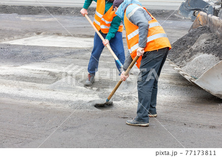 Two road workers rake old broken asphalt off the road and load it into a grader bucket. Two road workers rake old broken asphalt off the road and load it into a grader bucket. 77173811