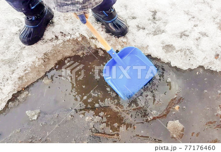 Close-up of a child's feet in rubber boots, a boy child happily playing with a shovel in a muddy puddle with snow and ice, in the early spring in the yard Close-up of a child's feet in rubber boots, a boy child happily playing with a shovel in a muddy puddle with snow and ice, in the early spring in the yard 77176460