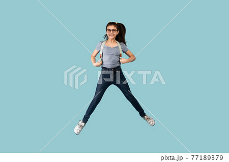 Excited Indian teen schoolgirl with backpack jumping up in air on blue studio background 77189379