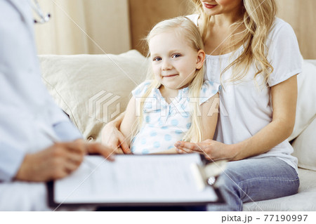 Doctor and patient. Pediatrician using clipboard while examining little girl with her mother at home. Happy cute caucasian child at medical exam. Medicine concept Doctor and patient. Pediatrician using clipboard while examining little girl with her mother at home. Happy cute caucasian child at medical exam. Medicine concept 77190997