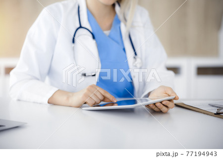 Unknown woman-doctor sitting in sunny clinic and using tablet pc, close-up. Data and best service in medicine Unknown woman-doctor sitting in sunny clinic and using tablet pc, close-up. Data and best service in medicine 77194943