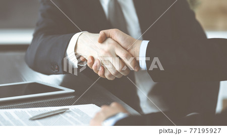 Unknown businessmen shaking hands above the glass desk in a modern office, close-up. Unknown business people at meeting. Teamwork, partnership and handshake concept 77195927