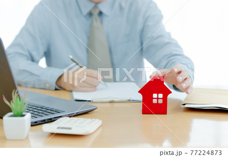 A House on desk envelope with man signing purchase documents in background. while hand complete the insurance policy, rental documents and loan document. concept guy buying new house 77224873