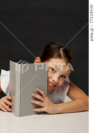little schoolgirl looks out from behind the book 77229540
