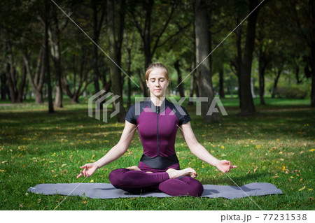 young beautiful slender blond woman in purple overall practice yoga in summer green park and demonstrates meditation and relaxation asana sukhasana 77231538