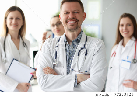 Successful smiling man doctor standing against background of colleagues in clinic Successful smiling man doctor standing against background of colleagues in clinic 77233729