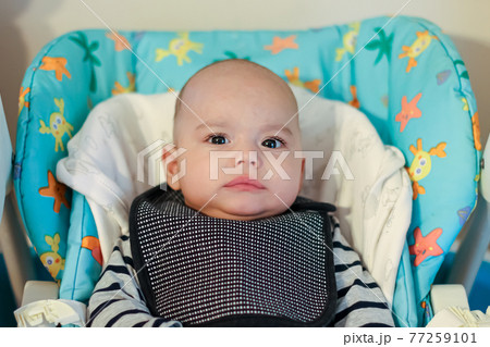 Baby in an apron sits in a highchair and waits to be fed. Serious baby looking to camera 77259101