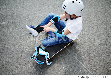 Sporty boy in safety helmet sitting on skateboard on playground asphalts and putting on protective knee pads 77268417