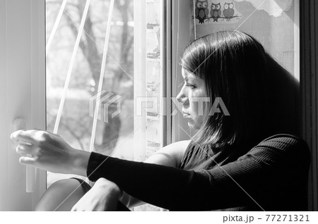 black and white image of a sad young pretty brunette woman in black clothes looking out the window 77271321