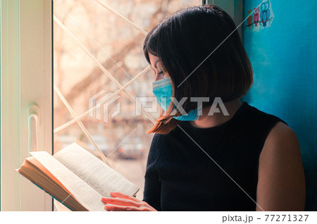 a dark-haired girl in a black dress sits in a surgical mask at the hospital window and reads a book a dark-haired girl in a black dress sits in a surgical mask at the hospital window and reads a book 77271327