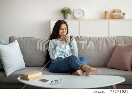 Full length of young Indian woman having coffee break on comfy sofa at home 77271925