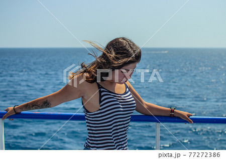 portrait of a pretty young girl with black hair with a tattoo on her arm in a striped tight T-shirt on a background of blue sea portrait of a pretty young girl with black hair with a tattoo on her arm in a striped tight T-shirt on a background of blue sea 77272386