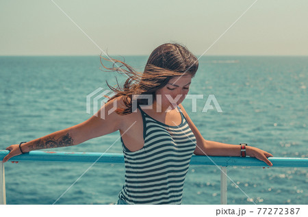 portrait of a beautiful young woman with black hair with a tattoo on her arm in a striped tight T-shirt on a background of blue sea portrait of a beautiful young woman with black hair with a tattoo on her arm in a striped tight T-shirt on a background of blue sea 77272387