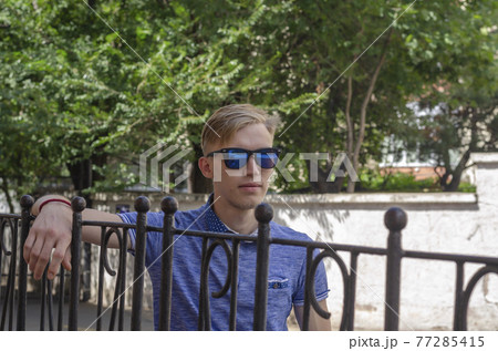 Handsome young man with blond hair stands at the metal fence. 77285415