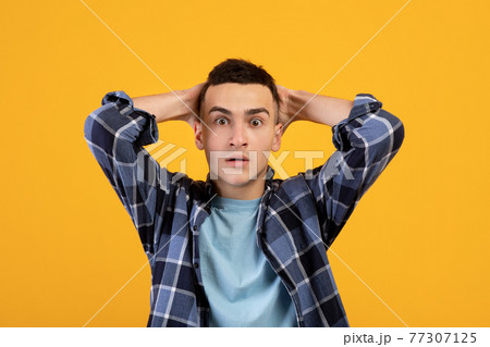 Emotional young man grabbing his head in shock, looking at camera on orange studio background. Human emotions Emotional young man grabbing his head in shock, looking at camera on orange studio background. Human emotions 77307125