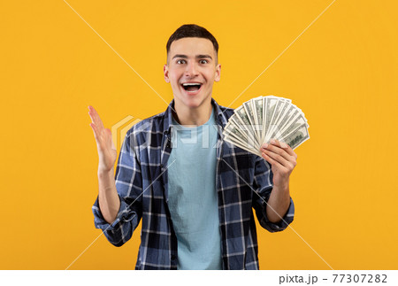 Handsome young man happy over winning big sum of money, holding dollar bills on orange studio background 77307282