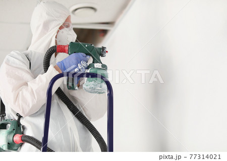 Male painter in protective suit is painting wall on stepladder with spray gun 77314021