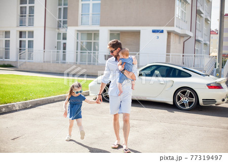 young dad walks with his two daughters on the background of a sports car 77319497