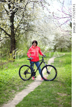 Beautiful woman on a bike in a blooming spring garden. Beautiful mature woman posing for the camera in a blooming spring garden. Beautiful woman on a bike in a blooming spring garden. Beautiful mature woman posing for the camera in a blooming spring garden. 77328541