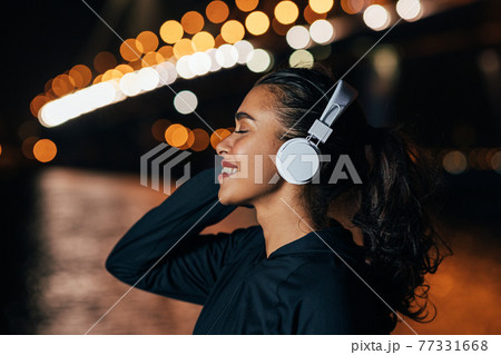 Side view of a fit woman in hooded shirt listening to music against night lights Side view of a fit woman in hooded shirt listening to music against night lights 77331668
