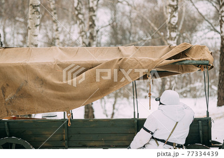 Hidden Re-enactor Dressed As German Wehrmacht Infantry Soldier In World War II Soldiers Sitting In Ambush Near Peasant Cart In Winter Forest And Looking At Old Army Binoculars. Historical Re-enactment 77334439