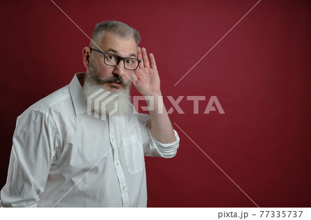 Mature bearded man dressed white shirt holds hand near ear, want to know information, red background Mature bearded man dressed white shirt holds hand near ear, want to know information, red background 77335737