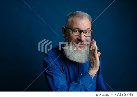 Bearded middle age man dressed blue shirt holds hand near mouth, secret talk concept, blue background Bearded middle age man dressed blue shirt holds hand near mouth, secret talk concept, blue background 77335748