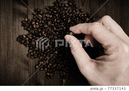 A man holds with his hand a coffee bean on a wooden background and a large amount of grain. Top view. 77337143