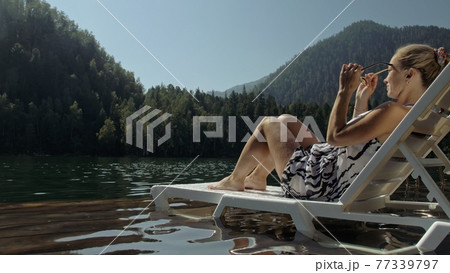 Woman lie on a sunbed in sunglasses and a boho silk shawl. Girl rest on a flood wood underwater pier. The pavement is covered with water in the lake. 77339797
