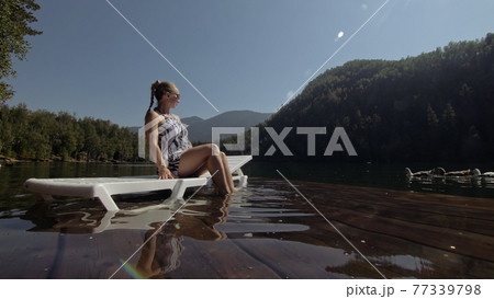 Woman lie on a sunbed in sunglasses and a boho silk shawl. Girl rest on a flood wood underwater pier. The pavement is covered with water in the lake. 77339798