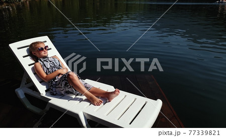 Kid girl lie on a sunbed in sunglasses and a boho silk shawl. Child rest on a flood wood underwater pier. The pavement is covered with water in lake. 77339821