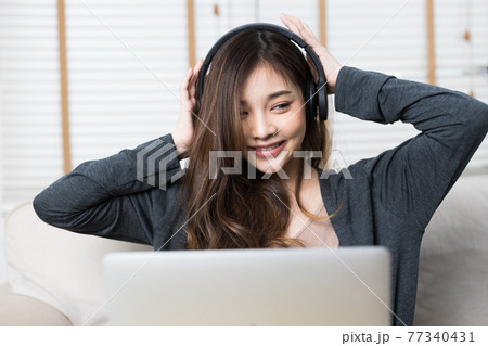 Cute young asian girl working on her lap top from the comfort of her home. She is in her sweater sitting on her couch. She is listening to music while smiling. Cute young asian girl working on her lap top from the comfort of her home. She is in her sweater sitting on her couch. She is listening to music while smiling. 77340431