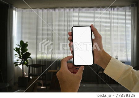 Man hands holding smart phone and blurred living room background. Blank screen for graphic display presentation montage. Man hands holding smart phone and blurred living room background. Blank screen for graphic display presentation montage. 77359328