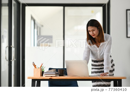 Beautiful woman working as secretary standing and look at her computer laptop over comfortable office as background. 77360410