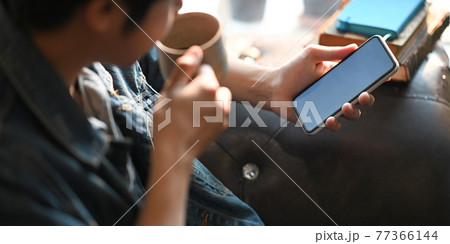 Cropped image of smart man drinking a hot coffee and holding a white blank screen smartphone in his hands while sitting/relaxing at the leather couch over comfortable sitting room as background. Cropped image of smart man drinking a hot coffee and holding a white blank screen smartphone in his hands while sitting/relaxing at the leather couch over comfortable sitting room as background. 77366144
