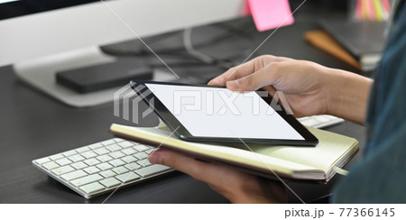Cropped image of smart man's hands holding a white blank screen computer tablet and notebook while sitting at the working desk over wireless keyboard and computer monitor as background. Cropped image of smart man's hands holding a white blank screen computer tablet and notebook while sitting at the working desk over wireless keyboard and computer monitor as background. 77366145