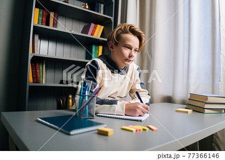 Low-angle view of thinking pupil boy thoughtful writing in notebook with pen sitting at desk near window. 77366146