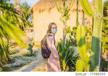 beautiful stylish young woman in pink dress in desert among the cacti, wearing a medical mask during COVID-19 coronavirus, traveling in Africa on safari, exploring nature, sunny summer, traveler on 77366294