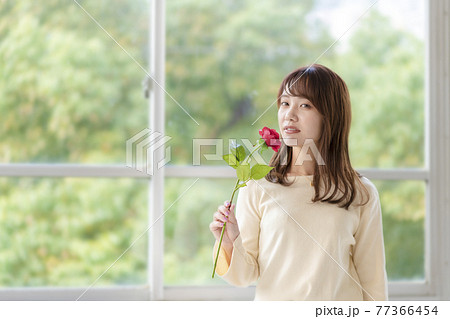 窓辺で薔薇の匂いを嗅ぐ若い女性 77366454