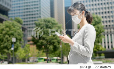 屋外でスマートフォンを操作するマスク姿のビジネスウーマン 屋外でスマートフォンを操作するマスク姿のビジネスウーマン 77368029