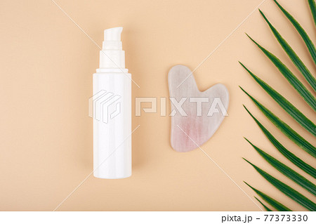 Top view of skin lotion, rose quartz heart shaped guasha massage stone and half of palm leaf on light beige background.  77373330