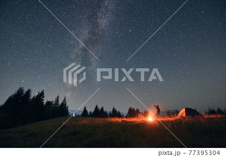 Male traveler standing near campfire and pointing at stars. 77395304