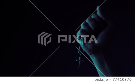 Praying with a rosary. hand of Catholic man with rosary on black background. Hands holding a silver rosary or cross pendant of Jesus Christ. Crucifix and hand on black background. Praying God concept 77410370