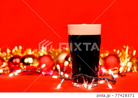 Dark ale beer in pint glass with christmas baubles tinsel and lights on red background 77428371