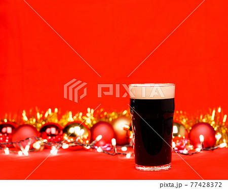 Dark ale beer in pint glass with christmas baubles tinsel and lights on red background Dark ale beer in pint glass with christmas baubles tinsel and lights on red background 77428372