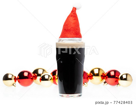 Dark ale beer in pint glass with christmas red hat and christmas baubles on white background Dark ale beer in pint glass with christmas red hat and christmas baubles on white background 77428403