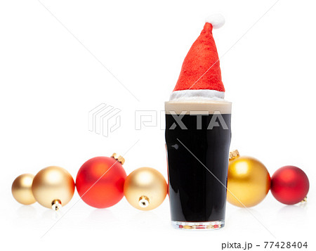 Dark ale beer in pint glass with christmas red hat and christmas baubles on white background Dark ale beer in pint glass with christmas red hat and christmas baubles on white background 77428404